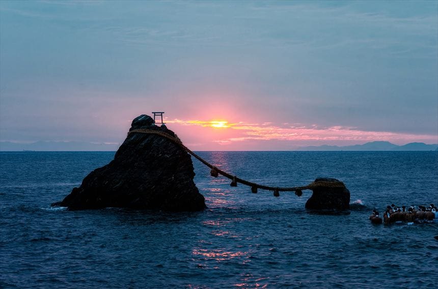 伊勢の夫婦岩の間に日が出たが富士山は雲で見えない。夏至祭で裸の男が海につかっている。