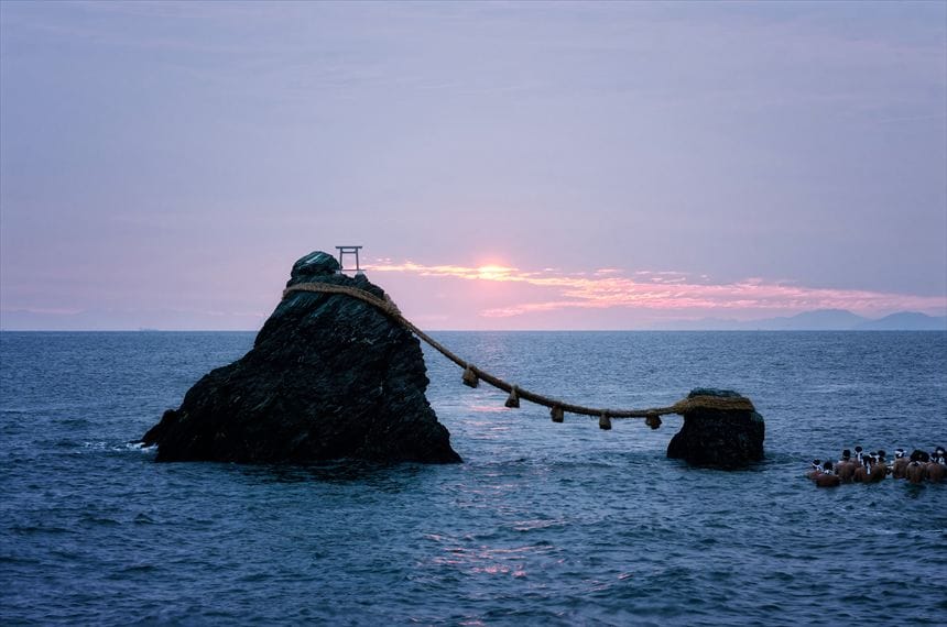 伊勢の夫婦岩の間に日が出たが富士山は雲で見えない。夏至祭で裸の男が海につかっている。