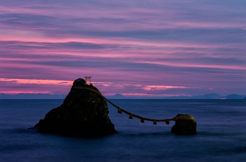 日の出前の伊勢の夫婦岩の間に富士山のシルエット。空は、異様な赤と黒の縞々に焼けている